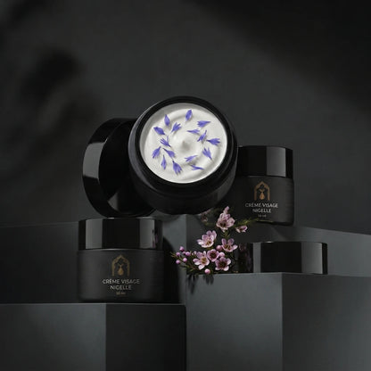 Deux pots de crème visage à la nigelle noirs, couvercles ouverts, crème blanche avec fleurs roses