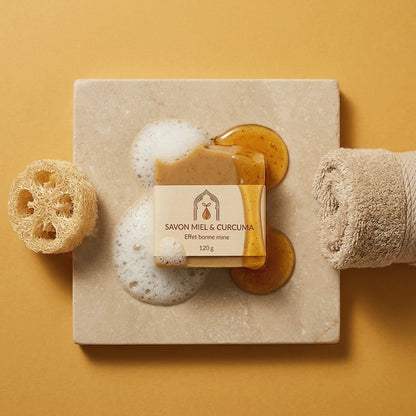 Savon miel et curcuma beige sur plaque, mousse blanche autour et gouttes ambrées.