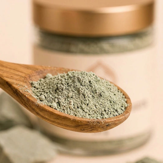 Cuillère en bois remplie de poudre d’argile verte, pot en verre visible derrière.