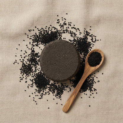 Barre de shampooing solide foncée et graines de nigelle sur une cuillère en bois.